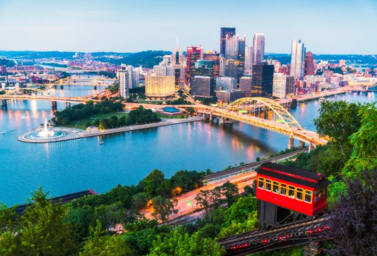 A panoramic view of a city skyline at dusk, showing tall buildings along the riverfront, multiple bridges crossing the water, and a large fountain at the point where two rivers meet. In the foreground, a bright red and yellow incline car travels up a steep, tree‑covered hillside.