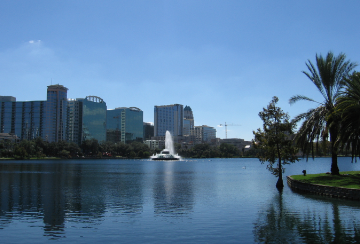 A calm city lake reflecting a clear blue sky, with modern high-rise buildings and a white fountain spraying water at the center; palm trees and grassy shoreline frame the right side of the scene.